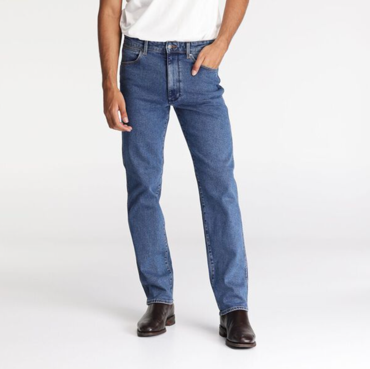 A person in Wrangler Classics Straight Jeans and a white t-shirt stands with one hand in their pocket, wearing brown leather shoes. The background is plain white.