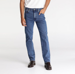 A person in Wrangler Classics Straight Jeans and a white t-shirt stands with one hand in their pocket, wearing brown leather shoes. The background is plain white.