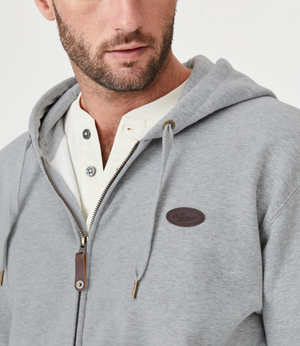 A man wears the RM Williams McKinlay Hooded Sweatshirt, a gray zip-up hoodie with a kangaroo pocket and small chest logo patch, layered over a white henley. The image captures his upper torso and part of his face.