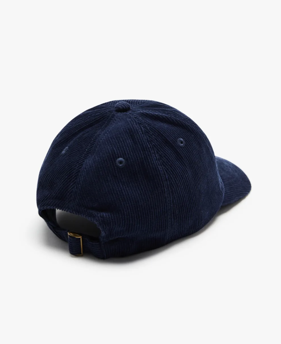 The RM Williams Corduroy Longhorn Cap, in navy blue cotton corduroy, is shown from the back on a white background. It has an adjustable strap with a brass buckle and an embroidered longhorn emblem, enhancing its rustic charm.