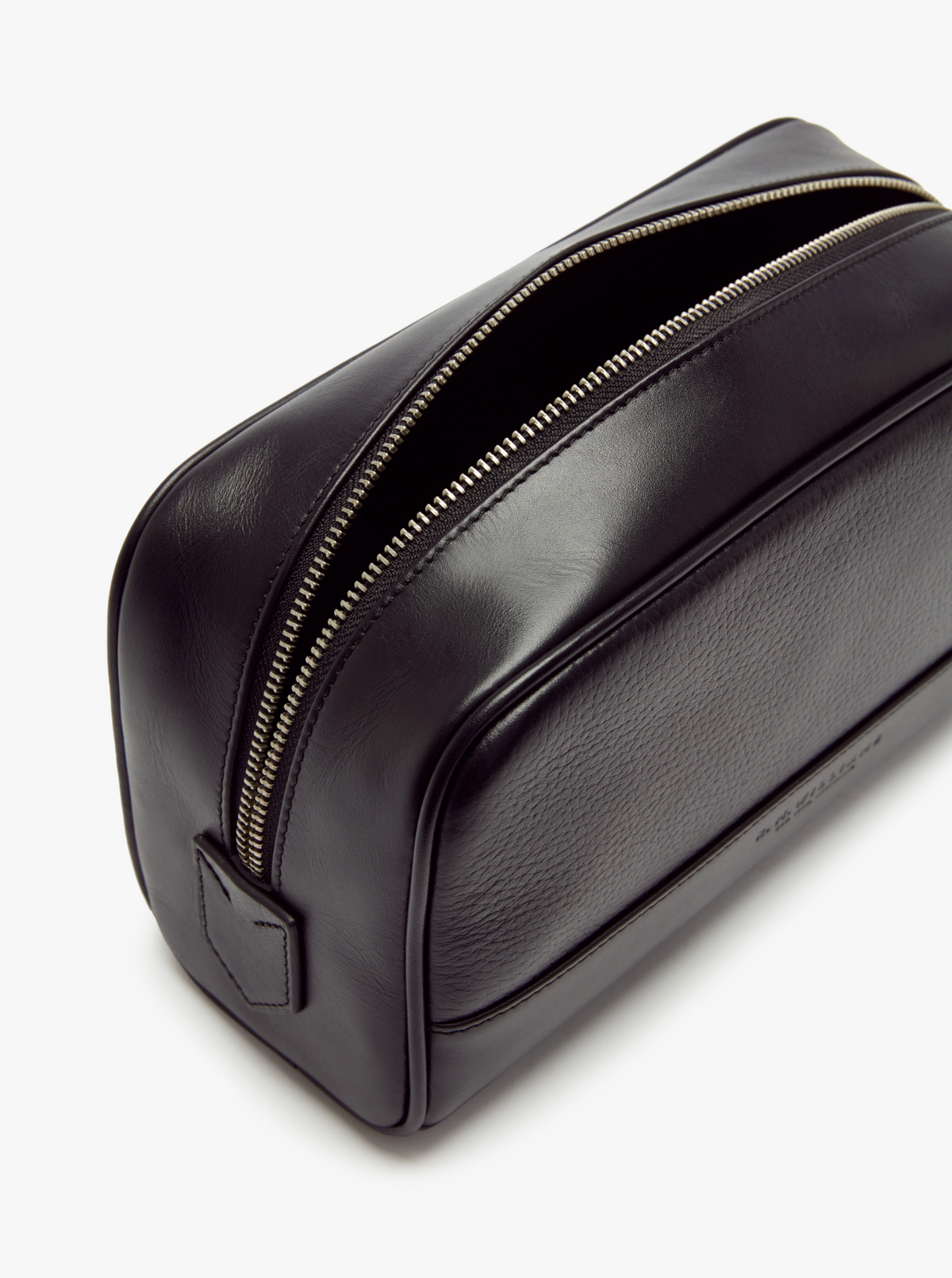The RM Williams City Washbag is a sleek, rectangular-shaped toiletry bag made from black full-grain leather. It features a single zipper along the top, partially open to showcase its spacious interior. The washbag has a smooth, slightly glossy finish with a small embossed logo on one end.
