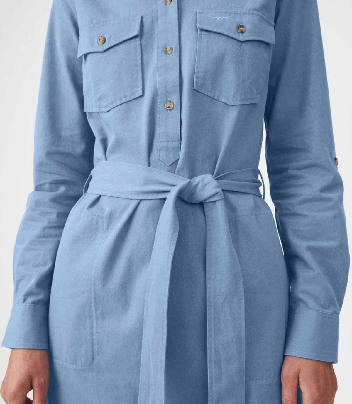 A person wears the RM Williams Adare Dress, a light blue chambray shirt dress with long sleeves, chest pockets, and a tie waistbelt. The image shows the dress cropped from shoulders to knees.