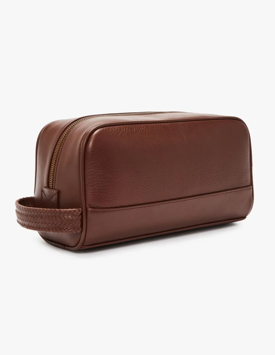 The RM Williams City Washbag is a brown leather toiletry bag featuring a gold zipper closure and a braided handle on one end. Crafted from full-grain leather, the bag boasts a simple, rectangular shape with a smooth and textured finish, set against a white background.