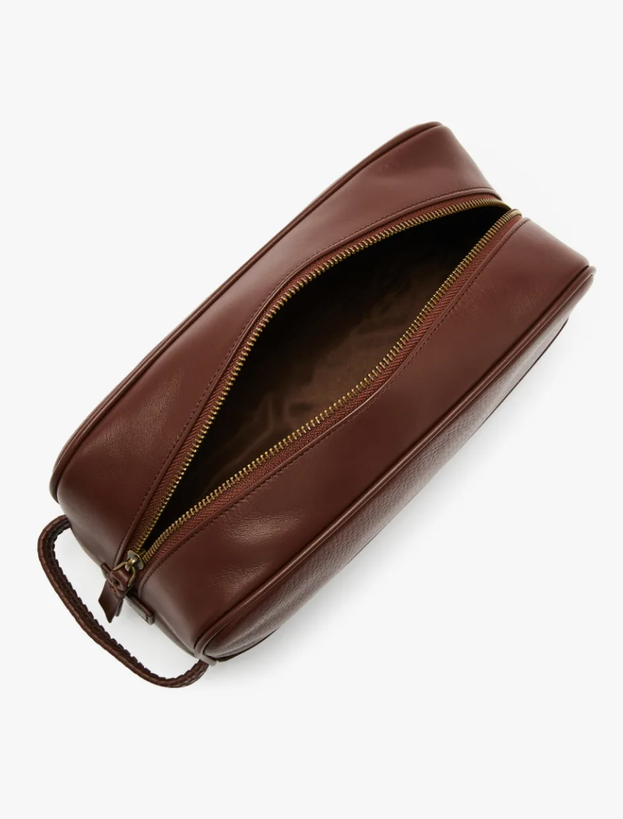 The RM Williams City Washbag, crafted from full grain brown leather, features a gold zipper partially open to reveal a patterned interior lining. With a handle on one side for easy carrying, this stylish toiletry bag is perfect for urban adventures.