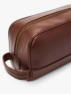 The RM Williams City Washbag is a brown leather accessory with a woven handle on the left side. It showcases a gold zipper running along the top and is crafted from high-quality, full-grain leather. The top panel has a smooth finish, while the sides have a slightly textured surface, exemplifying R.M.Williamsโ exceptional craftsmanship.