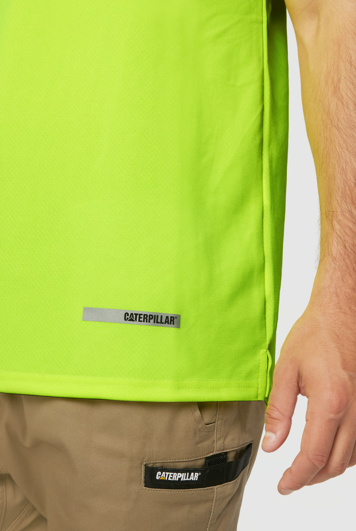 A person is wearing a CAT Hi Vis Performance Polo in bright lime green, with the CAT logo near the hem. The moisture control fabric shirt pairs with brown pants featuring a Caterpillar logo on the pocket, set against a plain white background.