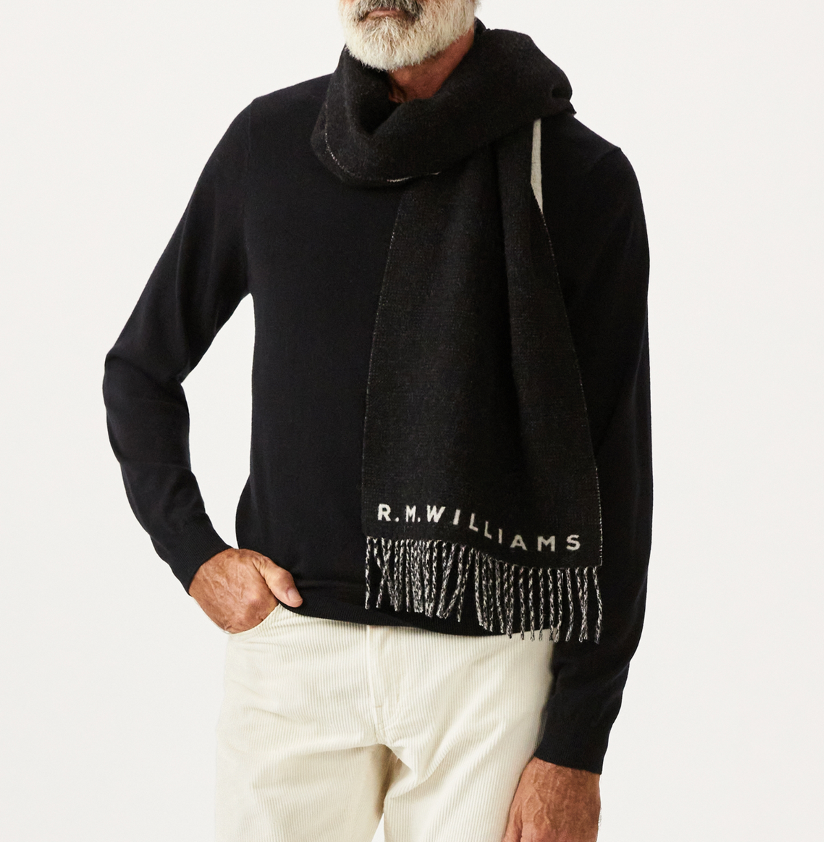 Draped in an RM Williams Logo Scarf, a person layers the stylish wool accessory featuring fringe detailing and a tassel trim over their black sweater and white pants, standing out against the plain white background.