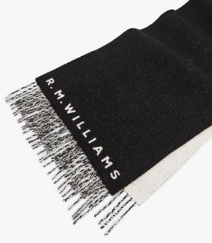 The RM Williams Logo Scarf features a black wool design with tassel trim and mixed black and white fringe, adorned with the "R.M. Williams" logo woven in white near the end for an elegant touch.