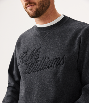 A bearded man wears an RM Williams Script Crew Neck jumper in a dark cotton blend, with the brand's name embroidered on it. A white shirt is visible at the collar against a plain light-colored background.