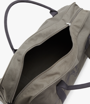 The RM Williams Nanga Canvas Bag is a grey cotton duffel-style bag with black handles, shown on a white background. Its zipper is partially open, exposing a dark, empty interior.