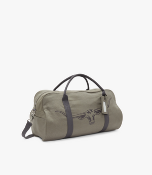 The RM Williams Nanga Canvas Bag is a khaki duffel-style cotton canvas bag with dark handles and strap. It features an embroidered longhorn skull design and has a hanging tag on one handle, set against a plain white background.
