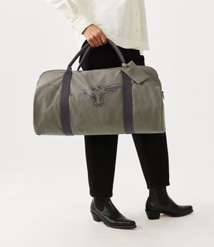 A person wearing a white shirt and black pants holds the gray RM Williams Nanga Canvas Bag with a cow design. The cotton canvas material pairs well with their black boots, all set against a plain white backdrop.