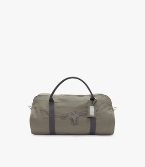 The RM Williams Nanga Canvas Bag is a gray cotton canvas duffel with black handles and a longhorn emblem on the front, featuring a rectangular luggage tag attached to one handle, set against a plain white background.