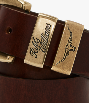 Here is a close-up of the RM Williams Drover Belt, crafted from vegetable-tanned leather, featuring an antique brass buckle engraved with "R.M. Williams" and an embossed longhorn cattle head design.