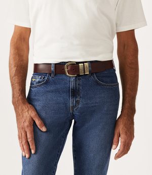 A person wearing a plain white t-shirt and blue jeans with the RM Williams Drover Belt, crafted from vegetable-tanned leather and featuring an antique brass buckle with two prongs. The individual's hands rest relaxed by their sides as they stand against a plain background.