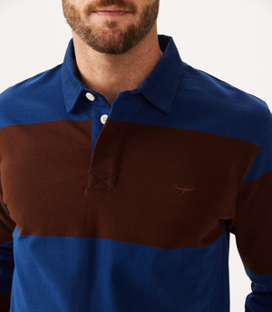 A person wearing the RM Williams Tweedale Rugby, a blue and brown heavyweight jersey rugby shirt with a small Longhorn chest embroidery and partially buttoned collar. Only the upper torso and part of their bearded face are visible.