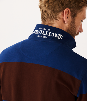 A person with light brown hair is facing away, wearing the RM Williams Tweedale Rugby shirt, featuring a blue and brown collar embroidered with "AUSTRALIA, R.M. WILLIAMS, Est. 1932" in white. A subtle Longhorn embroidery adorns the plain white background for a unique touch.