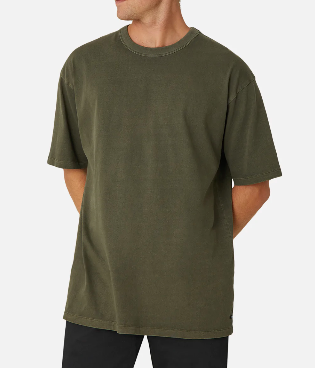 A person models the Industrie The Del Sur Tee—an oversized olive green t-shirt with a relaxed crew neckline and short sleeves. Paired with black pants, they stand against a white background, hands behind their back, highlighting its style and comfort.