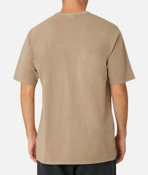 A person is shown from the back wearing Industrie The Del Sur Tee, a plain beige 100% cotton casual T-shirt with an oversized fit for comfort, paired with dark pants. A small tag is visible at the collar, and the backdrop is a plain white.