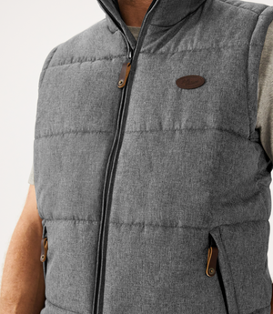 A person is wearing the RM Williams Patterson Creek Vest, a gray quilted puffer with zip and buttoned pockets, an adjustable drawstring hem, and a small brown leather chest patch over a light gray shirt against a plain white background.