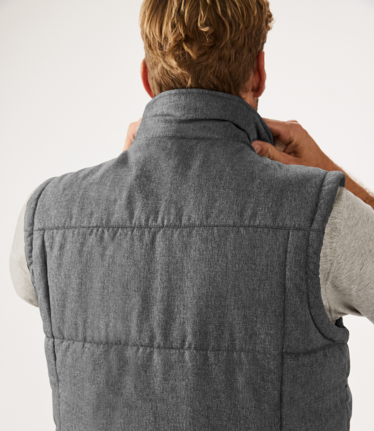 A person with short blond hair, viewed from behind, adjusts the collar of a gray RM Williams Patterson Creek Vest over a light gray shirt, against a plain white background.