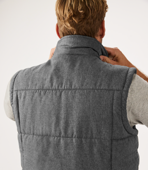 A person with short blond hair, viewed from behind, adjusts the collar of a gray RM Williams Patterson Creek Vest over a light gray shirt, against a plain white background.