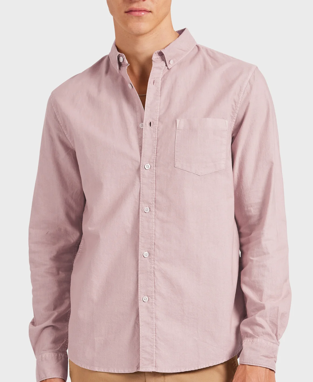 A person is wearing the Academy Brand Vintage Oxford Shirt in light pink, featuring a refined point collar and crafted from 100% Cotton Oxford. The shirt has a relaxed fit, is buttoned to the collar, and is paired with khaki pants against a plain white background.