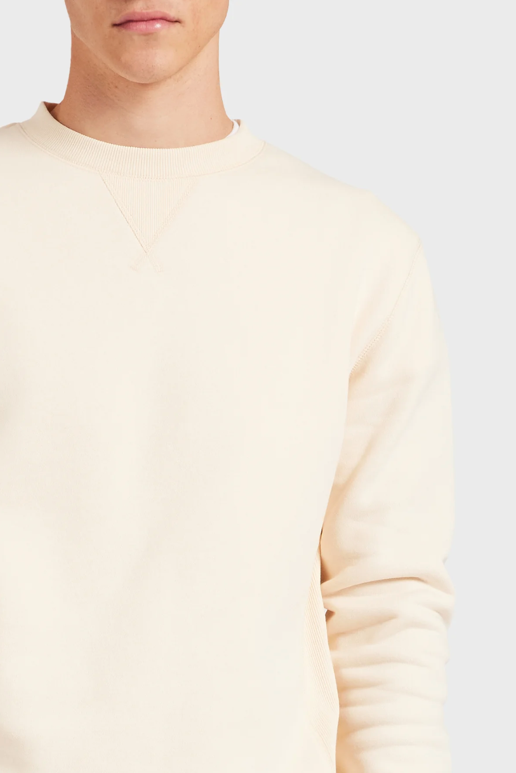 A person models the Academy Brand Crew Sweat—a plain, cream-colored, relaxed-fit sweatshirt—against a light gray backdrop, with only their upper body and lower face visible.