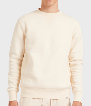 A person wears the Academy Brand Crew Sweat, a plain cream-colored brushed fleece sweatshirt with ribbed cuffs and hem, paired with matching relaxed-fit sweatpants, all set against a light grey background.