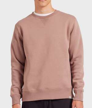 A person wears the Academy Brand Crew Sweat—a relaxed fit, long-sleeve, light brown brushed fleece crewneck—over a white T-shirt and dark pants. The image is cropped at the mouth and thighs.