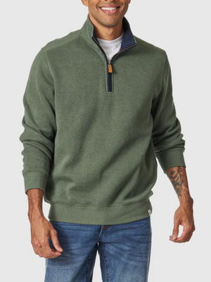 A man stands smiling in the Blazer Heather Half Zip Sweat, a green cotton-polyester quarter-zip sweater with a brown zipper pull and ribbed texture, worn over a white T-shirt and blue jeans. His hands rest relaxed at his sides.