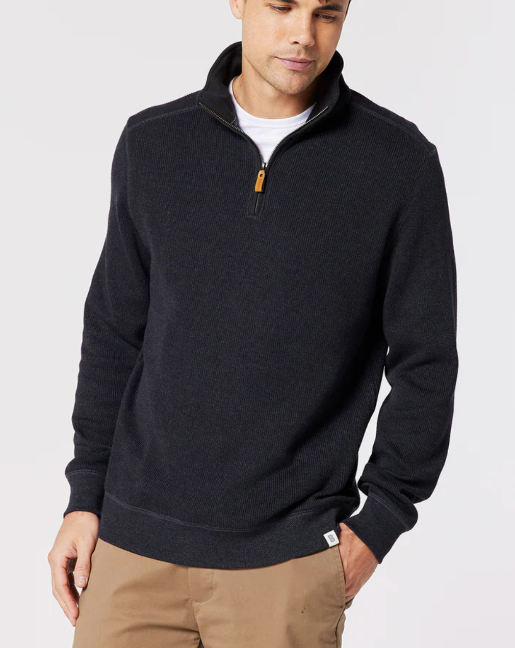 A man models the Blazer Heather Half Zip Sweat over a white shirt and light brown pants, standing against a plain light background with one hand in his pocket and looking slightly down.