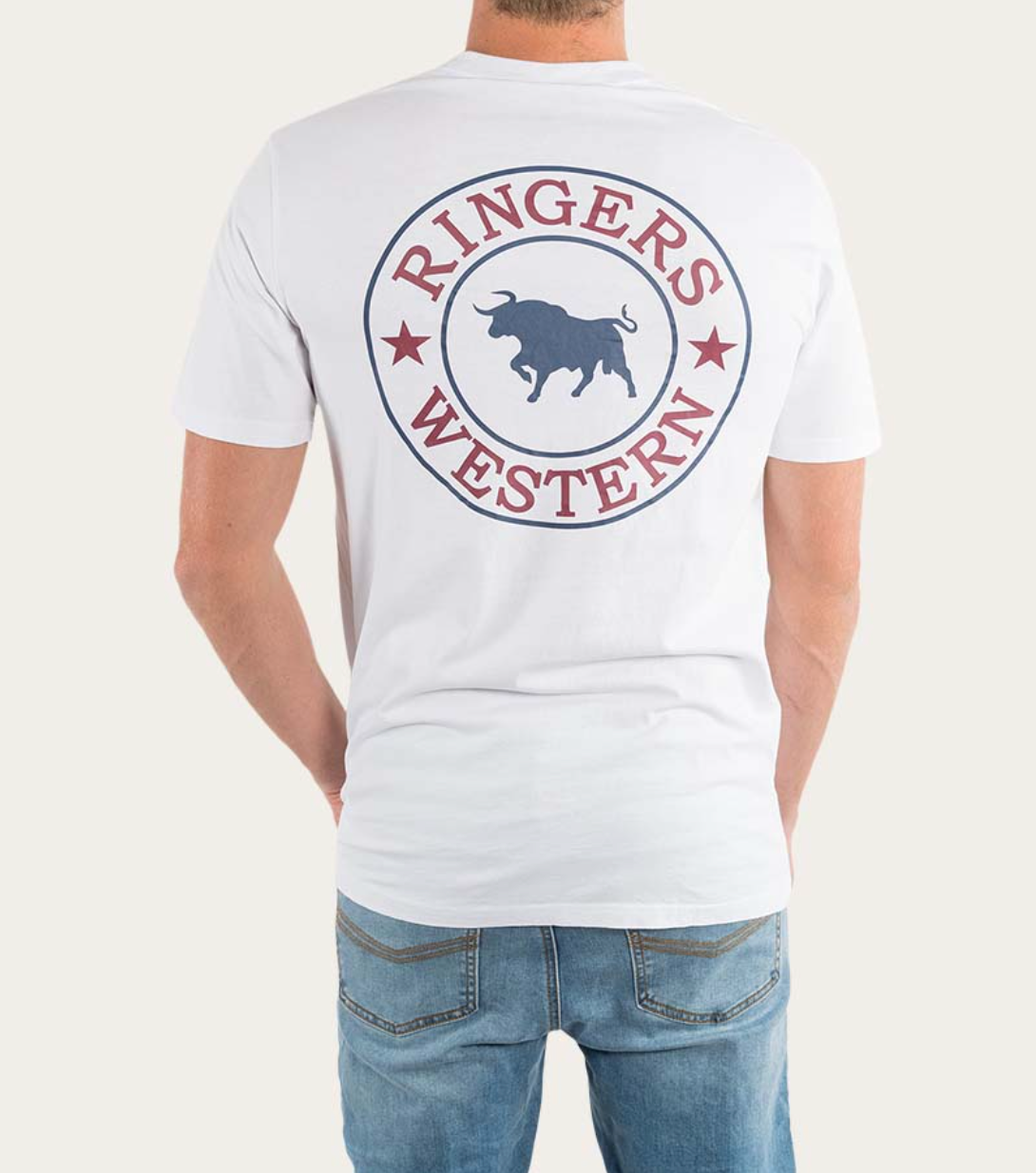 A person wearing the Ringers Western Signature Bull Classic T-Shirt, which is a comfortable white tee featuring a large circular logo on the back with a blue silhouette of a bull and the text "RINGERS WESTERN" in red. Made from pre-shrunk cotton, this classic fit tee pairs perfectly with their blue jeans as they face away from the camera.