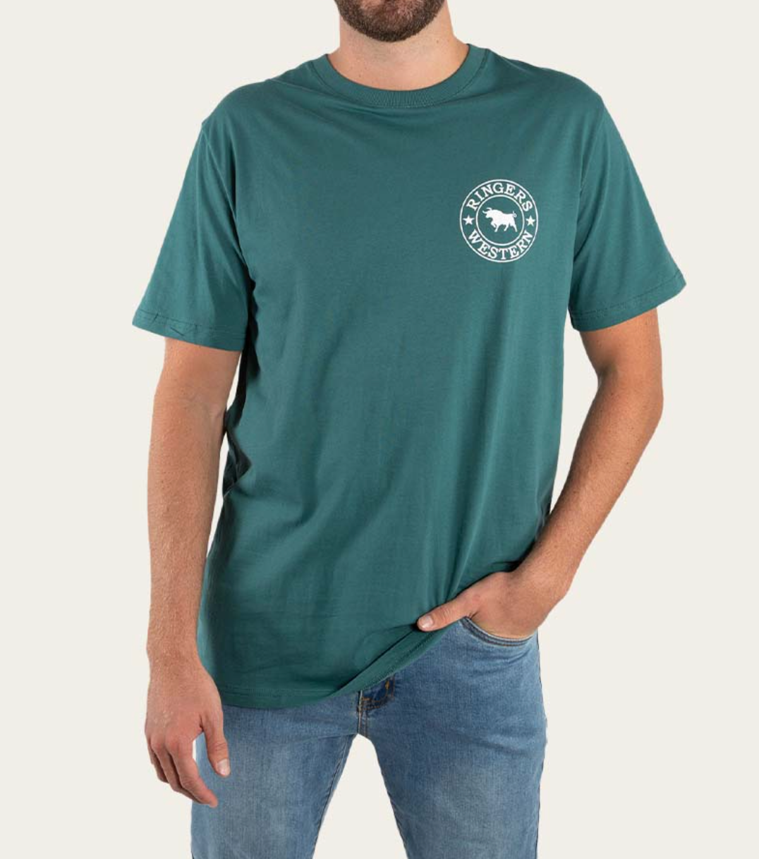 A person wears a teal Ringers Western Signature Bull Classic T-Shirt, made of pre-shrunk cotton, with the logo—a circle with text and a bull silhouette—on the upper left. They pair it with blue jeans and have one hand in their pocket against a plain white background.