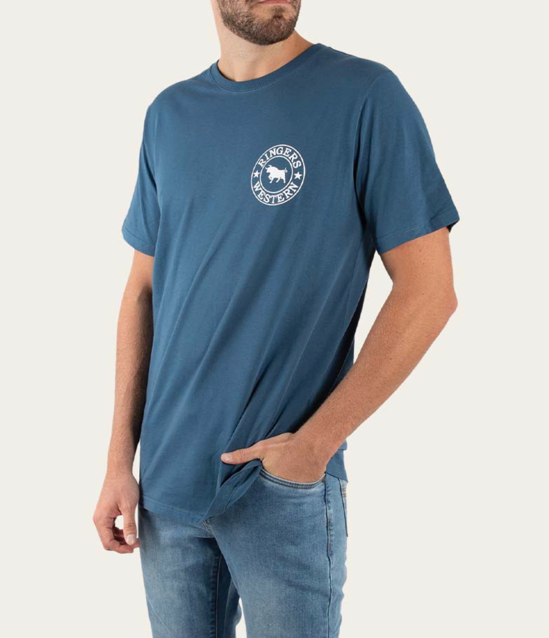 A man stands confidently with one hand in his pocket, wearing a Ringers Western Signature Bull Classic blue short-sleeve tee made of pre-shrunk cotton featuring a small circular logo on the chest, paired with light blue jeans.