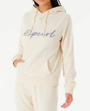 The person is wearing a comfortable beige Rip Curl Script II Hood with "Ripcurl" in blue on the front, featuring a cozy fleece lining and a front pocket where one hand rests casually against the plain white background.