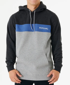 A person wears the Rip Curl Undertow Panel Hood, featuring a dark gray upper section, blue stripe with "Ripcurl" text, and light gray lower section. This comfy fleece-lined hoodie has white drawstrings and a front pocket. They stand against a plain white background.