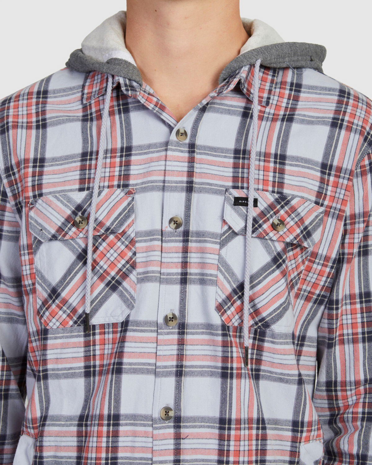 A close-up shows someone in a Rip Curl Ranchero Flannel Shirt, featuring red, white, and blue plaid patterns. Made from soft cotton, it has two chest pockets and a grey hood with drawstrings; the person's face isn't visible.