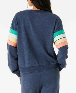 A person with long hair is wearing the Rip Curl Day Break Crew, a relaxed fit blue sweatshirt and pants. The crew neck sweatshirt features colorful sleeve stripes in green, turquoise, peach, and beige. The person is facing away from the camera.