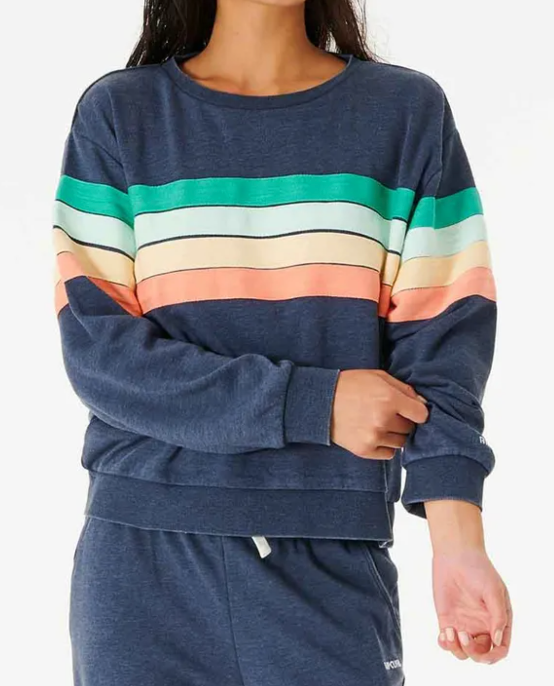 A person in a relaxed fit, blue Rip Curl Day Break Crew sweatshirt featuring horizontal stripes in green, white, peach, and yellow adjusts their sleeve. They're wearing matching blue joggers against a plain white background.