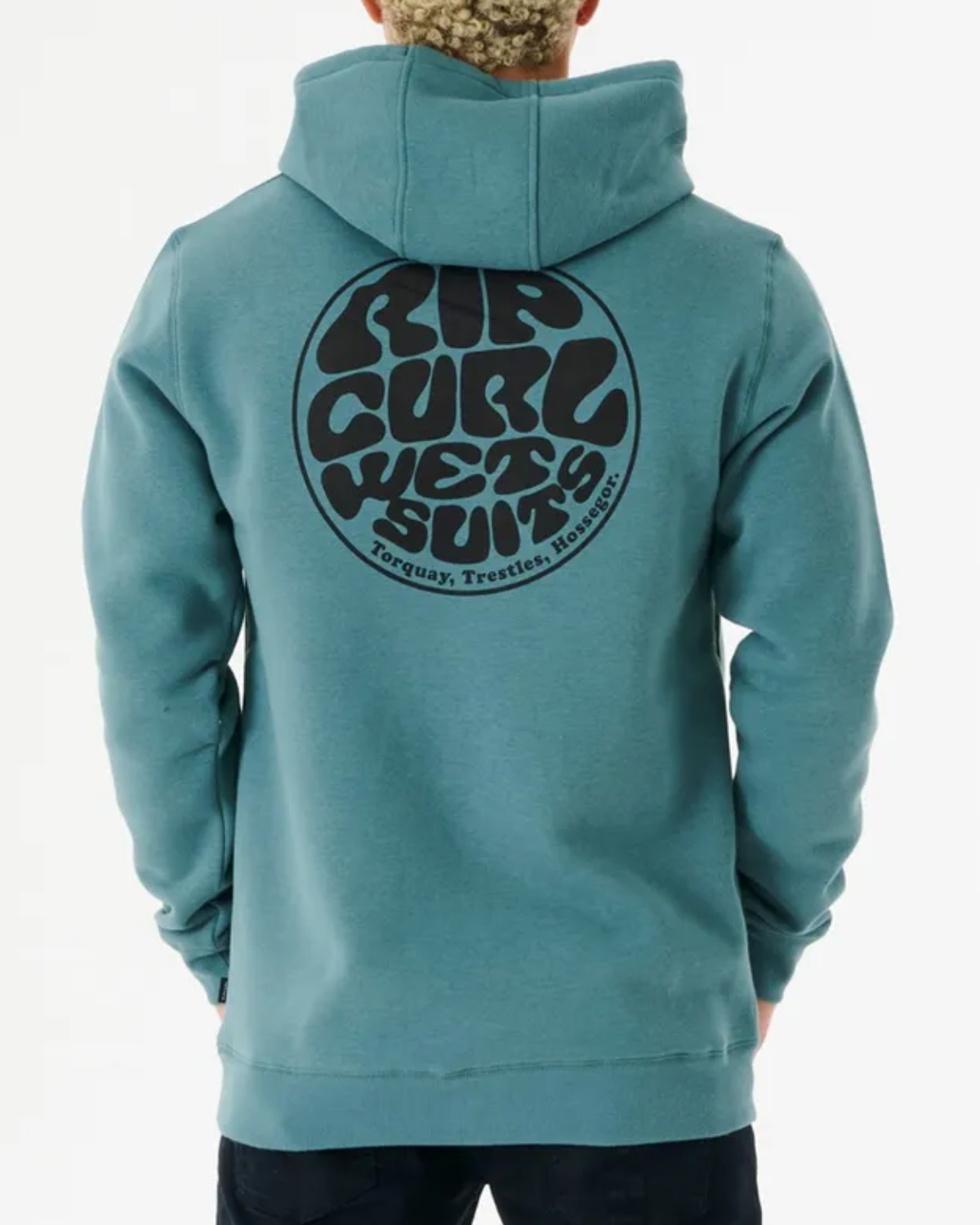 A person with curly hair is wearing a blue Rip Curl Wetsuit Icon Hood, showcasing a bold circular logo on the back that reads "Rip Curl Wet Suits, Torquay, Trestles, Hossegor." Made from organic cotton, the hoodie blends style and sustainability as they face away from the camera.