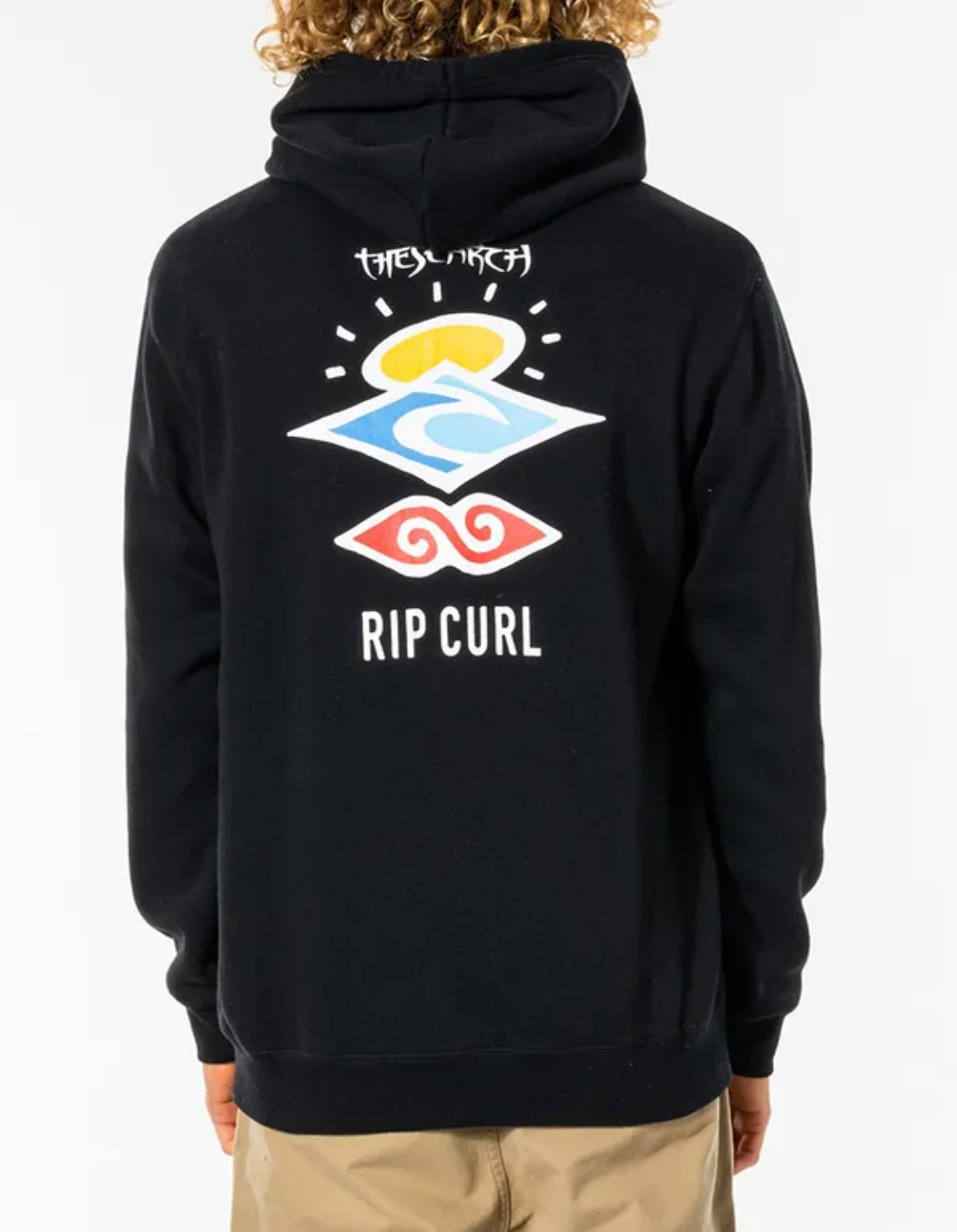 Back view of a person with curly blonde hair wearing the Rip Curl Search Icon Hood, a comfortable black casual hoodie. The hoodie, made from soft fleece lining and Organic Brushed Cotton, features a colorful wave design along with the words "THE SEARCH" and "RIP CURL" printed on the back. The person is also wearing beige pants.