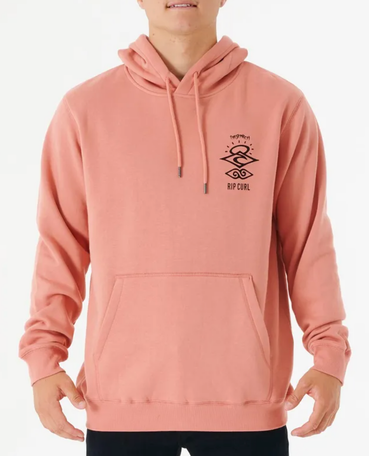 A person wearing a Rip Curl Search Icon Hood in peach, featuring front graphics. Made from organic brushed cotton with a soft fleece lining for comfort and style, it includes a front pocket and drawstrings, elegantly paired with black pants on a plain white background.