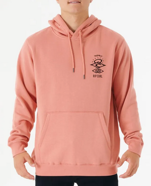 A person wearing a Rip Curl Search Icon Hood in peach, featuring front graphics. Made from organic brushed cotton with a soft fleece lining for comfort and style, it includes a front pocket and drawstrings, elegantly paired with black pants on a plain white background.