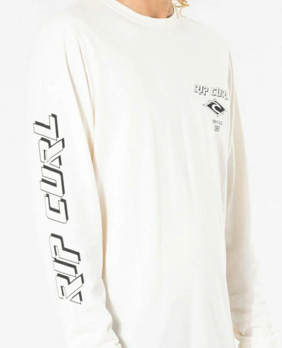 Wearing the Rip Curl Fade Out Icon Long Sleeve Tee, a person showcases "RIP CURL" in bold along the right sleeve. This white, sustainable organic cotton shirt features a small chest logo and text in sleek black, offering minimalistic branding suited for any casual outing.