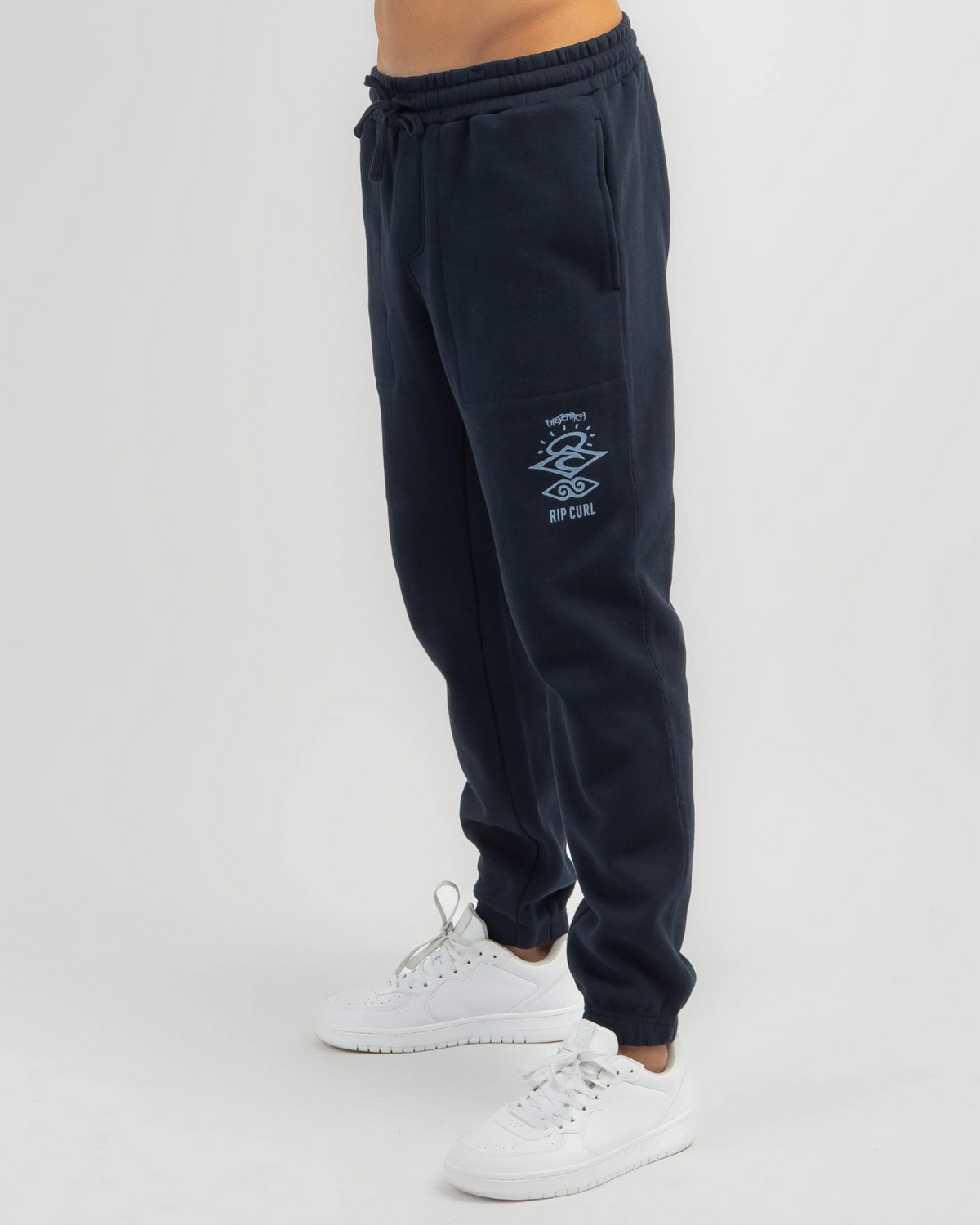 A person wears Rip Curl Search Icon Track Pant in navy blue featuring a small white graphic on one leg, paired with sleek white sneakers. Made from Organic Brushed Cotton, the joggers stand out against the plain white background.