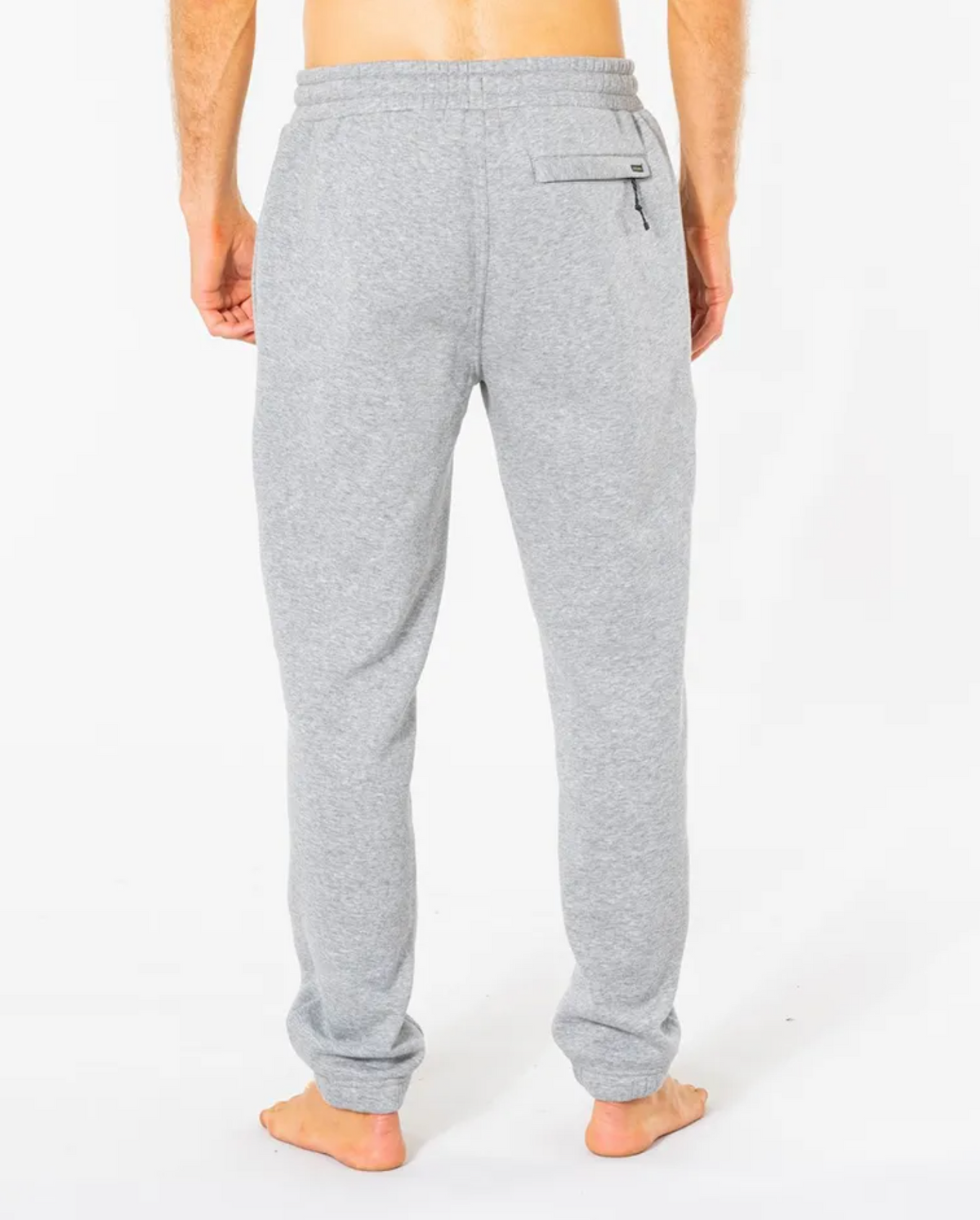 A person is seen barefoot from behind against a white background, wearing light gray Rip Curl Search Icon Track Pants. These comfy pants are crafted from organic brushed cotton and feature an elastic waistband with a back right pocket.