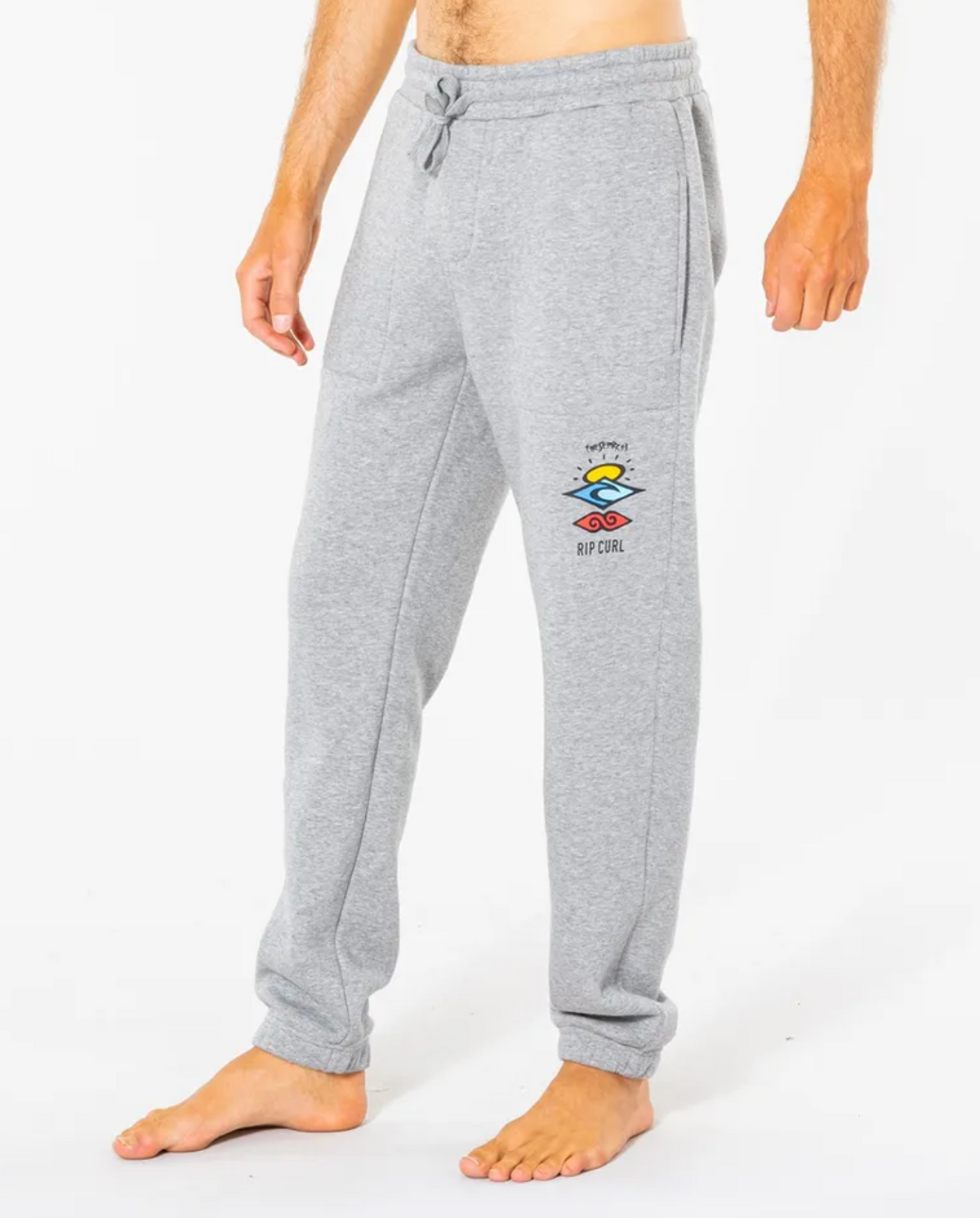 A person in light gray Rip Curl Search Icon Track Pants with a colorful logo on the left thigh, made of organic brushed cotton, featuring an elastic drawstring waist, stands barefoot against a plain white background.