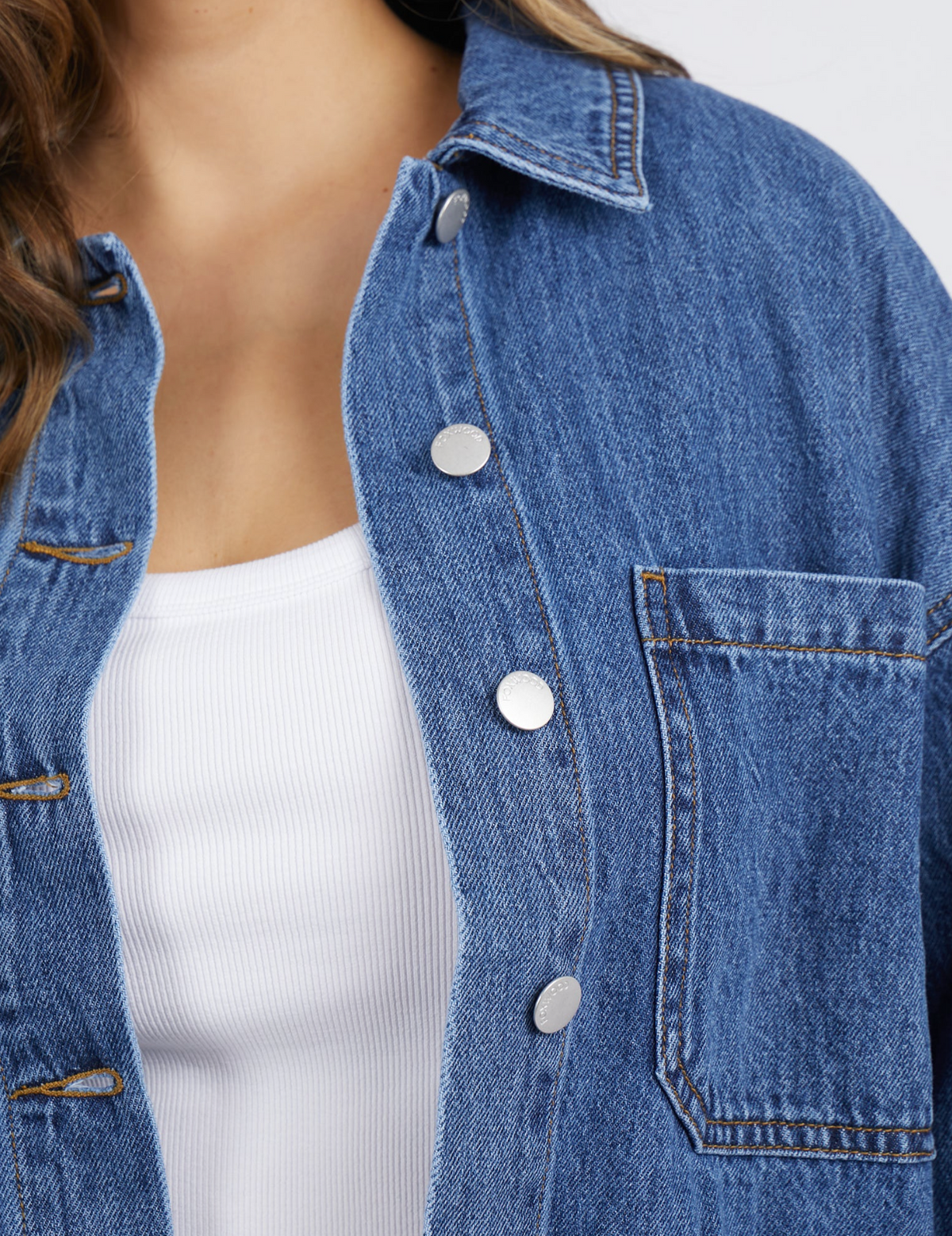 A close-up shows a person in a Foxwood Joelle Denim Shacket, featuring a relaxed fit made from 100% cotton in blue denim. It's worn over a white ribbed shirt, partially buttoned, with a left-side pocket against a plain white background.