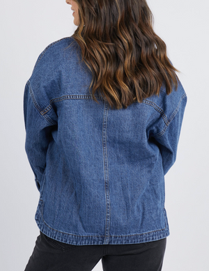 A woman with wavy brown hair, facing away, wears the Foxwood Joelle Denim Shacket and black pants against a gray background. The relaxed fit adds comfort and style while showcasing the shacket's 100% cotton material.